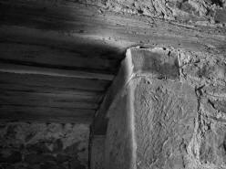 PAŘÍŽOV: povalový strop z dřevěných hraněných trámů nad chodbičkou v západní zdi a kamenné ostění s dřevěným překladem portálu vedoucího ke schodišti v tloušťce zdi. Na stěnách místnosti z rulových desek jsou viditelné podřezávané spáry. V pozadí se nachází zazdívka románského portálu (foto M. Falta 2010).